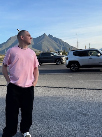 Lucho Miranda, de cabello corto, camiseta rosa y pantalón negro, sonríe ligeramente con las manos en los bolsillos. Al fondo, montañas y tráfico en una carretera