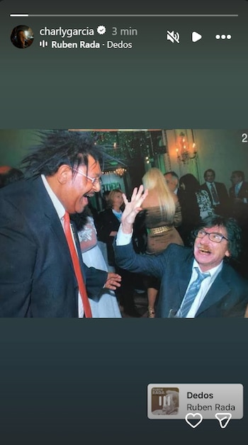 Fotografía de Rubén Rada de perfil sonriendo junto a Charly García, quien levanta la mano derecha en un evento