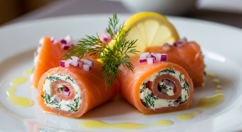 Rollitos de salmón ahumado con relleno cremoso y eneldo, decorados con cebolla morada picada y una rama de eneldo, con una rodaja de limón en un plato blanco.