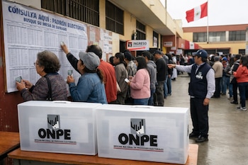 En una jornada electoral, personas en Perú buscan su mesa de votación en carteles. Hay ánforas de la ONPE en primer plano y un oficial señalando. Bandera de Perú al fondo.