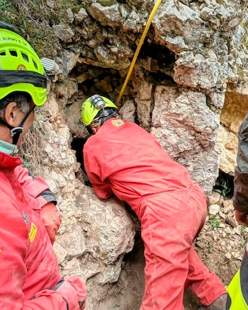 Rescatistas vestidos con monos rojos y cascos amarillos inspeccionan la entrada de una estrecha cavidad entre rocas