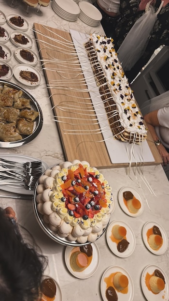 Una mesa con una tarta decorada con cintas blancas, una pavlova con frutas frescas y varios platos con flan y dulce de leche