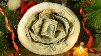 Vista superior de un gorro de Navidad lleno de billetes de dólares estadounidenses, entre ramas de pino, cintas rojas y una vela.