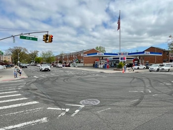 Vista de una intersección con semáforo y señal de 69 St, una gasolinera Mobil con bandera estadounidense, y múltiples marcas de derrape en el asfalto