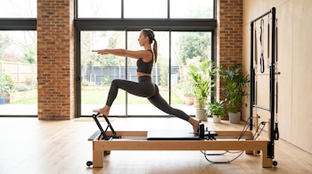 Mujer con ropa deportiva gris oscuro realiza una estocada en un reformer de madera, con los brazos extendidos y mirada al frente, en un estudio con ventanales.
