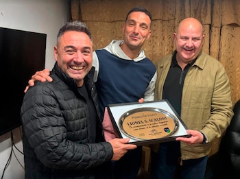 Tres hombres sonrientes sostienen una placa conmemorativa que lleva el nombre Lionel S. Scaloni. Detrás, cortinas marrones y una pantalla oscura