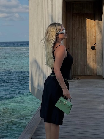 Wanda Nara de perfil, con cabello rubio, gafas de sol, top y falda negros, y un bolso verde, de pie en una pasarela de madera sobre agua azul