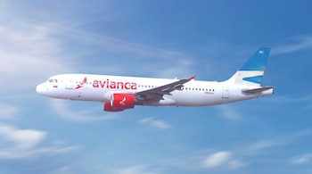 Un avión blanco de Avianca con motores rojos y cola azul y blanca vuela en un cielo azul claro con nubes tenues.