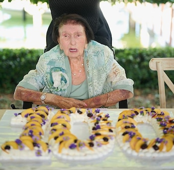 Una mujer mayor sentada detrás de una mesa con tres pasteles que forman el número 100, decorados con frutas y flores moradas