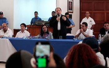 Un hombre con traje oscuro y camisa blanca habla por un micrófono en un podio con una mesa azul, flanqueado por personas sentadas y de pie