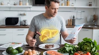 Un hombre en una cocina come carne, con un esquema de hígado amarillo sobre su abdomen. En la mesa hay huevos, aguacates, salmón, lechuga y un libro.