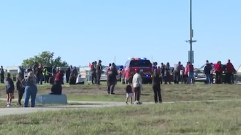 Personas congregadas en un terreno de césped junto a vehículos policiales y de emergencia con luces encendidas bajo la luz del día