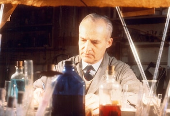 Un hombre con camisa, corbata y chaqueta escribe detrás de frascos de vidrio con líquidos de colores e instrumental de laboratorio