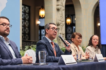 Jonathan Menkos habla en un micrófono mientras está sentado en un panel junto a tres personas. De fondo se ven arcos de piedra y lámparas decorativas