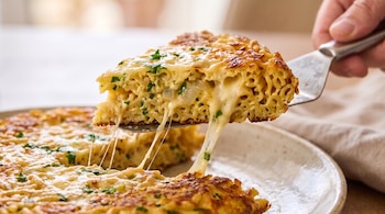 Una mano sostiene una espátula que levanta una porción de tortilla de pasta con queso derretido sobre un plato.