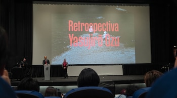 Un auditorio con un escenario y una pantalla grande. En la pantalla se lee 'Retrospectiva Yasujiro Ozu'. Tres personas en el escenario. Público en asientos.