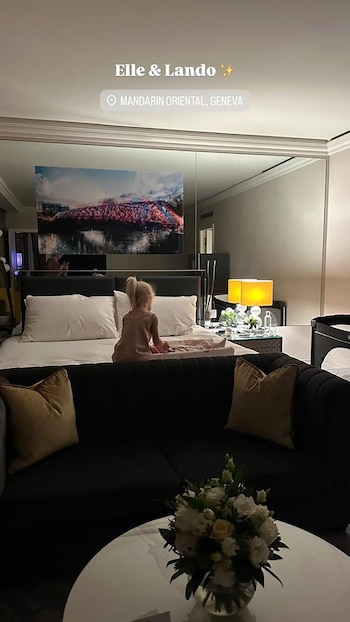 Vista de una habitación de hotel con una niña sentada de espaldas en una cama grande, un sofá con cojines, una cuna y un arreglo floral en una mesa