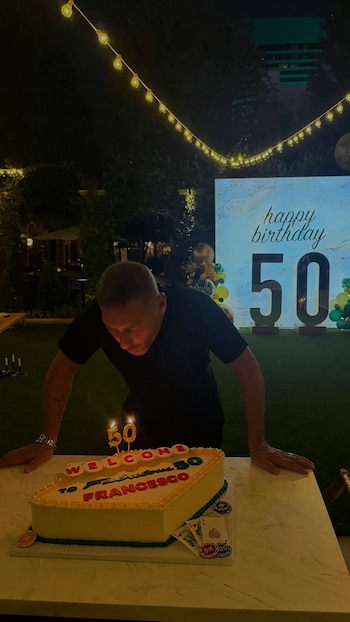 Un hombre se inclina sobre un pastel decorado con cartas de póquer y el número 50 encendido frente a un cartel de cumpleaños