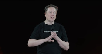 Elon Musk vestido con una camiseta negra con la palabra Roadster, sosteniendo un micrófono frente a un fondo oscuro