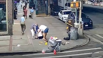 Tres personas agachadas en una acera junto a varios objetos en el suelo, vehículos estacionados y un semáforo en Nueva York