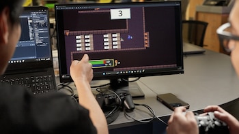 Dos personas frente a pantallas; una señala un monitor con un videojuego pixelado de retos matemáticos, la otra sostiene un control de videojuegos en un escritorio