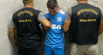 Tres hombres, uno con camiseta celeste de fútbol siendo sujetado por dos policías de espaldas con chalecos "Antinarcóticos Policía" frente a una pared texturada
