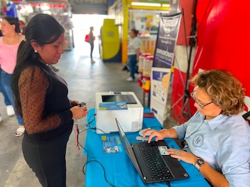Una mujer sentada junto a una computadora e impresora revisa el documento de una ciudadana que está de pie frente al escritorio