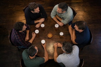 Vista superior de seis hombres sentados a una mesa redonda con vasos de cerveza y un pequeño plato con aperitivos.