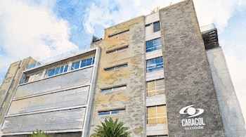 Fachada de edificio de varios pisos con ventanas rectangulares, paredes texturizadas, letrero de Caracol Televisión. Cielo azul y nubes.