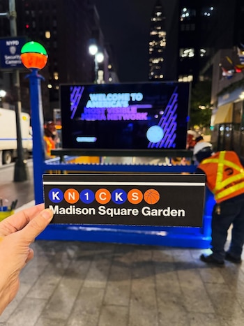 Una mano sostiene un cartel negro con el logotipo de los Knicks y "Madison Square Garden" frente a una pantalla digital y una estructura azul en la noche de Nueva York