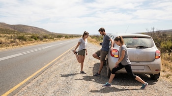 Dos mujeres y un hombre se estiran las piernas junto a un automóvil claro estacionado al costado de una carretera asfaltada en un paisaje árido con colinas.