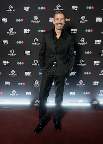 Un hombre vestido con traje negro sonríe frente a un panel negro con logotipos de MasterChef Celebrity y HBO Max