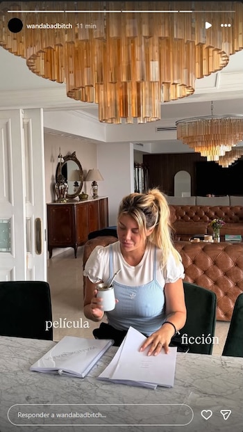 Wanda Nara, con cabello rubio recogido, sentada a una mesa de mármol, leyendo papeles y bebiendo mate en un salón elegante con candelabros