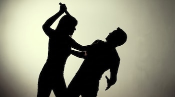 Siluetas oscuras de una mujer levantando un objeto sobre un hombre que se inclina hacia atrás, sobre un fondo liso y claro.