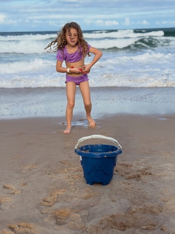 Una niña con traje de baño color violeta está de pie en la orilla del mar con una pala roja, frente a un balde azul. Detrás se observan olas