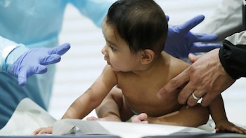 Un bebé está en una mesa de examen médico. Una mano enguantada azul se ve a la izquierda y una mano sin guante toca el hombro del bebé a la derecha