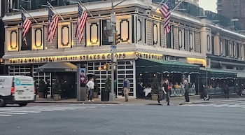 Exterior da Smith & Wollensky em Nova York ao entardecer, com bandeiras americanas, mesas ao ar livre, pedestres na calçada e carros na rua