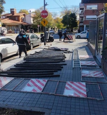 Un agente de policía camina junto a barras metálicas y barreras de construcción en una acera, con vehículos y una señal de PARE en una intersección