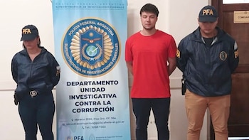 Ignacio Veltri, un joven vestido de rojo, de pie entre dos agentes de la PFA uniformados frente a un banner de la Policía Federal Argentina
