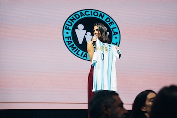 Una mujer sostiene un micrófono y una camiseta de fútbol a rayas celestes y blancas frente a una pantalla con un logotipo circular