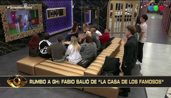Un grupo de personas sentadas en un sofá dorado, mirando una pantalla grande que muestra a dos hombres en un set de televisión verde