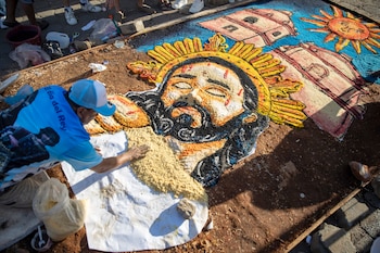 Un hombre con gorra azul crea una alfombra de aserrín colorida en el suelo, con diseños de un sol radiante y dos estructuras arquitectónicas coloniales, en una calle empedrada