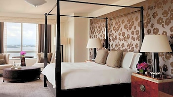 Una habitación de hotel de lujo con cama de cuatro postes, ropa de cama blanca, dos sillones, mesa de centro, y un gran ventanal con vistas al mar