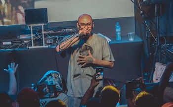 Hombre calvo con gafas y tatuajes, camiseta estampada, canta en micrófono. Detrás hay equipo de DJ y un portátil. Público con teléfonos lo graba