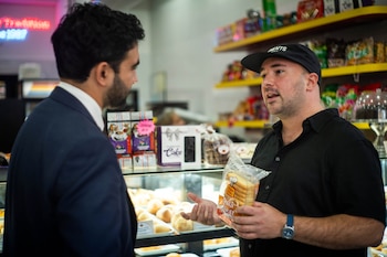 Un hombre en traje escucha a un comerciante con gorra que sostiene un paquete de pan dentro de un establecimiento con mostrador y estantes