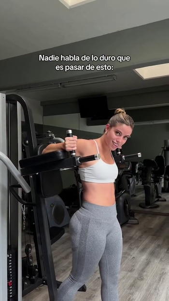 Una mujer en un gimnasio usa una máquina de ejercicio, viste top blanco y leggings grises. Tiene el pelo recogido y mira sonriendo a cámara. Fondo de espejos y equipos