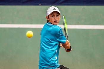 Un joven tenista, Ignacio Buse, vestido con una camiseta azul y gorra blanca, golpea una pelota de tenis amarilla con una raqueta verde en una cancha de tenis
