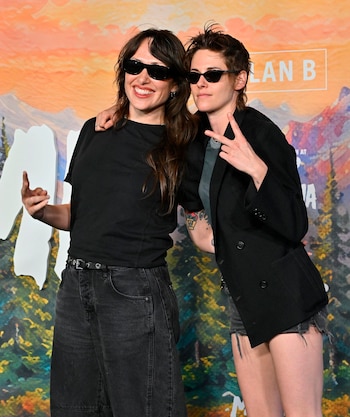 Kristen Stewart y otra mujer posan con gafas de sol oscuras frente a un fondo con montañas. Kristen Stewart viste saco negro y hace un signo de paz