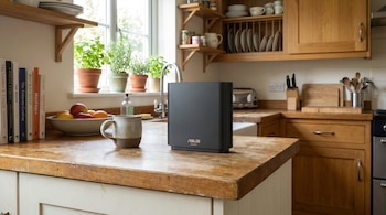 Router ASUS ZenWiFi negro vertical sobre una encimera de madera en una cocina con ventanas, plantas en macetas, libros, fregadero y armarios de madera.