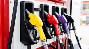 Cuatro boquillas de combustible de colores amarillo, rojo, morado y negro, con sus mangueras negras, cuelgan en un dispensador blanco y rojo.